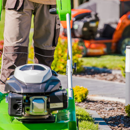 LandscapingA person in brown pants operates a green lawnmower on a sunny day. Vibrant green grass and colorful shrubs are visible, with another mower blurred in the background.