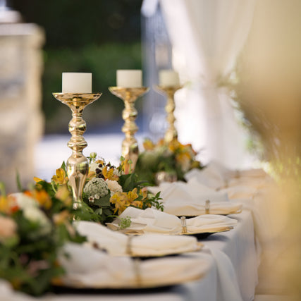 Party & EventElegant table setting with tall gold candleholders and white candles, surrounded by lush floral arrangements with yellow and orange blooms, exuding a festive, sophisticated ambiance.