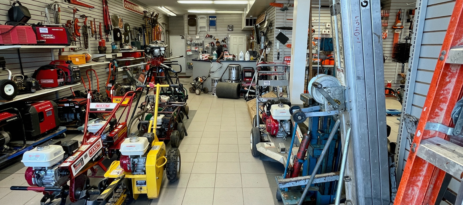 A hardware store aisle filled with various tools and equipment, including lawnmowers and ladders. Shelves on the sides display hand tools, creating a busy, functional atmosphere.